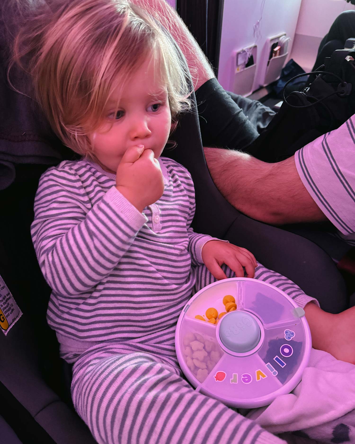 Blonde toddler in striped pajamas eating snacks out of snack box on airplane