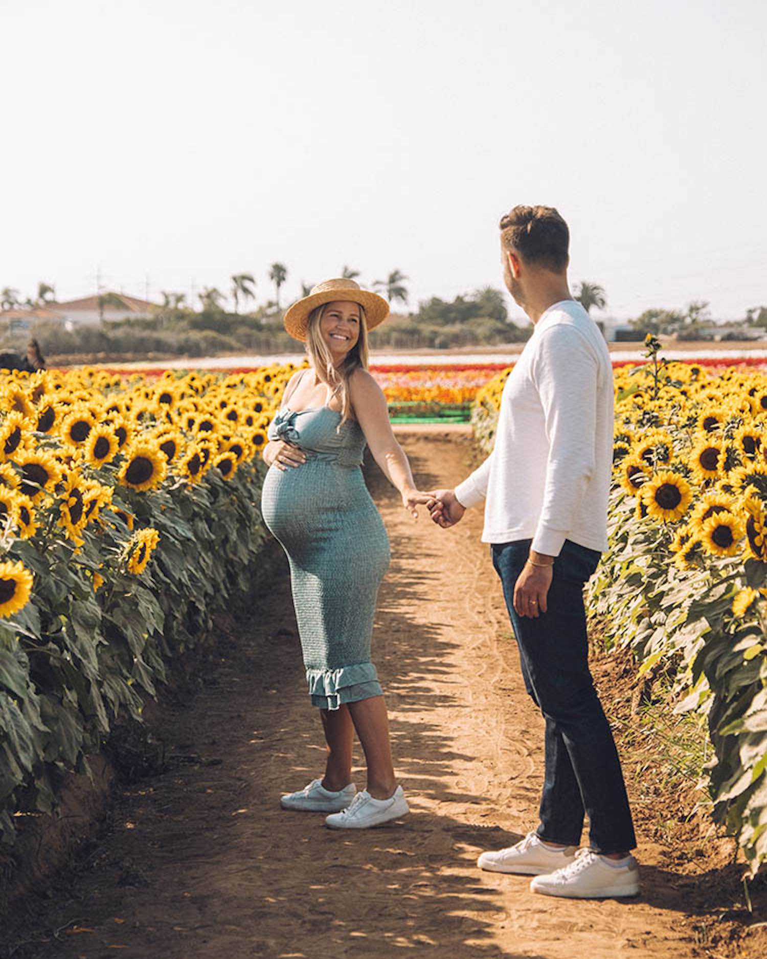 Pregnant woman in green body con dress wandering sunflower fields as she looks back at man whose hand she's holding as he wears white long sleeve and jeans