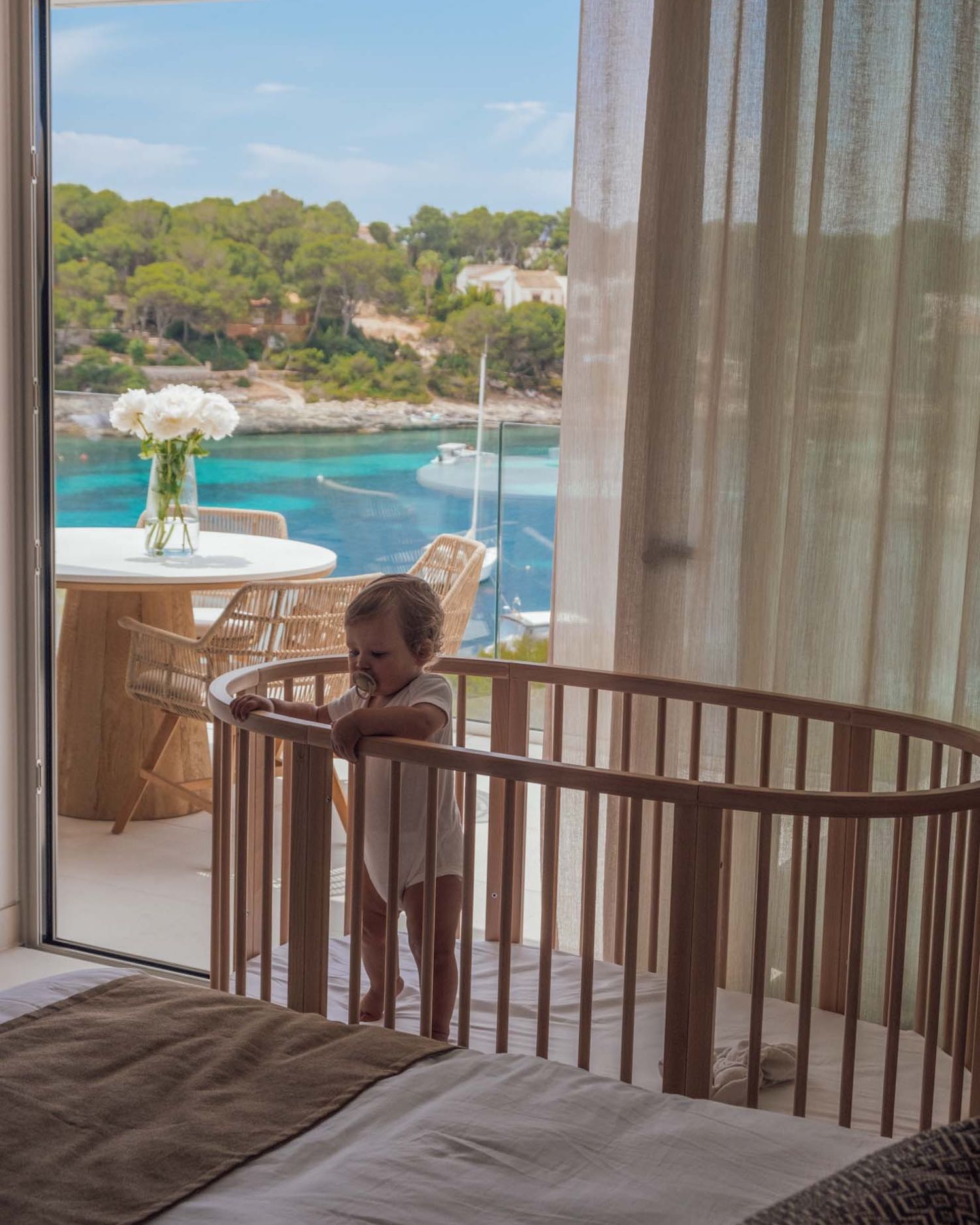 Baby standing in beige wooden crib next to edge of bed with curtains behind and view of outdoor white table and view of ocean