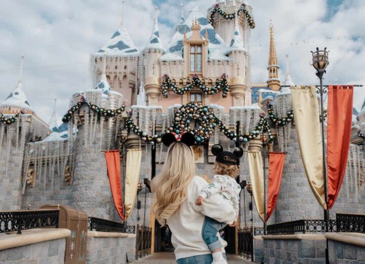 back of blonde woman with Minnie mouse ears holding blonde toddler boy in Mickey Mouse cap facing Sleeping Beauty Castle at Disneyland