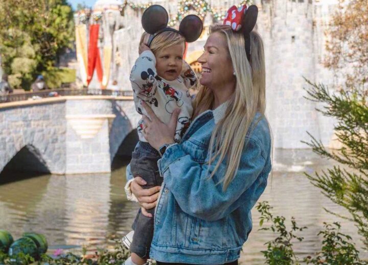 Blonde woman smiling in jean jacket and minnie mouse ears holding blonde toddler in mickey ears and mickey sweatshirt in front of Disneyland Sleeping Beauty castle