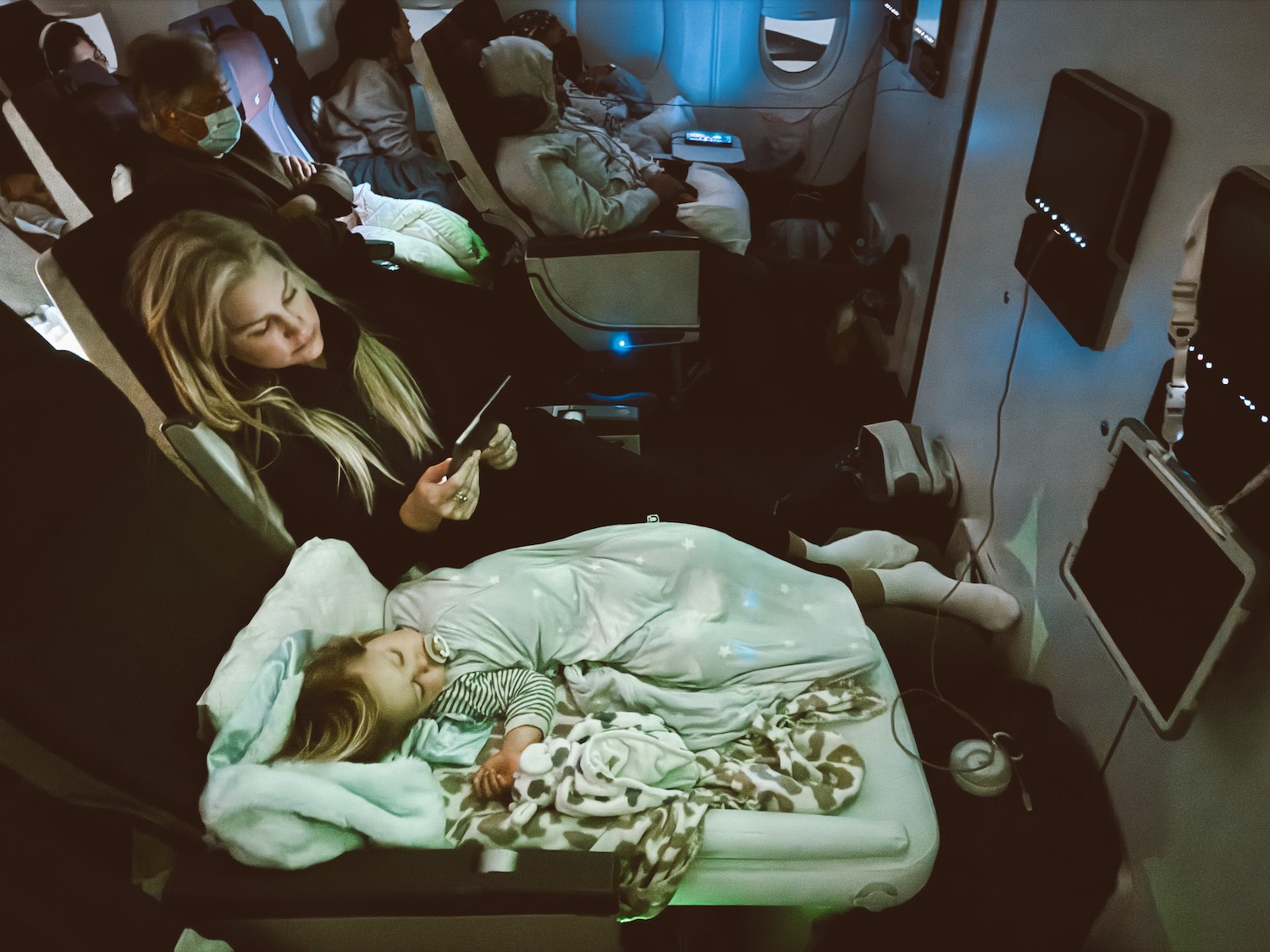 baby sleeping on portable inflatable mattress on airplane with blonde mom on tablet next to baby