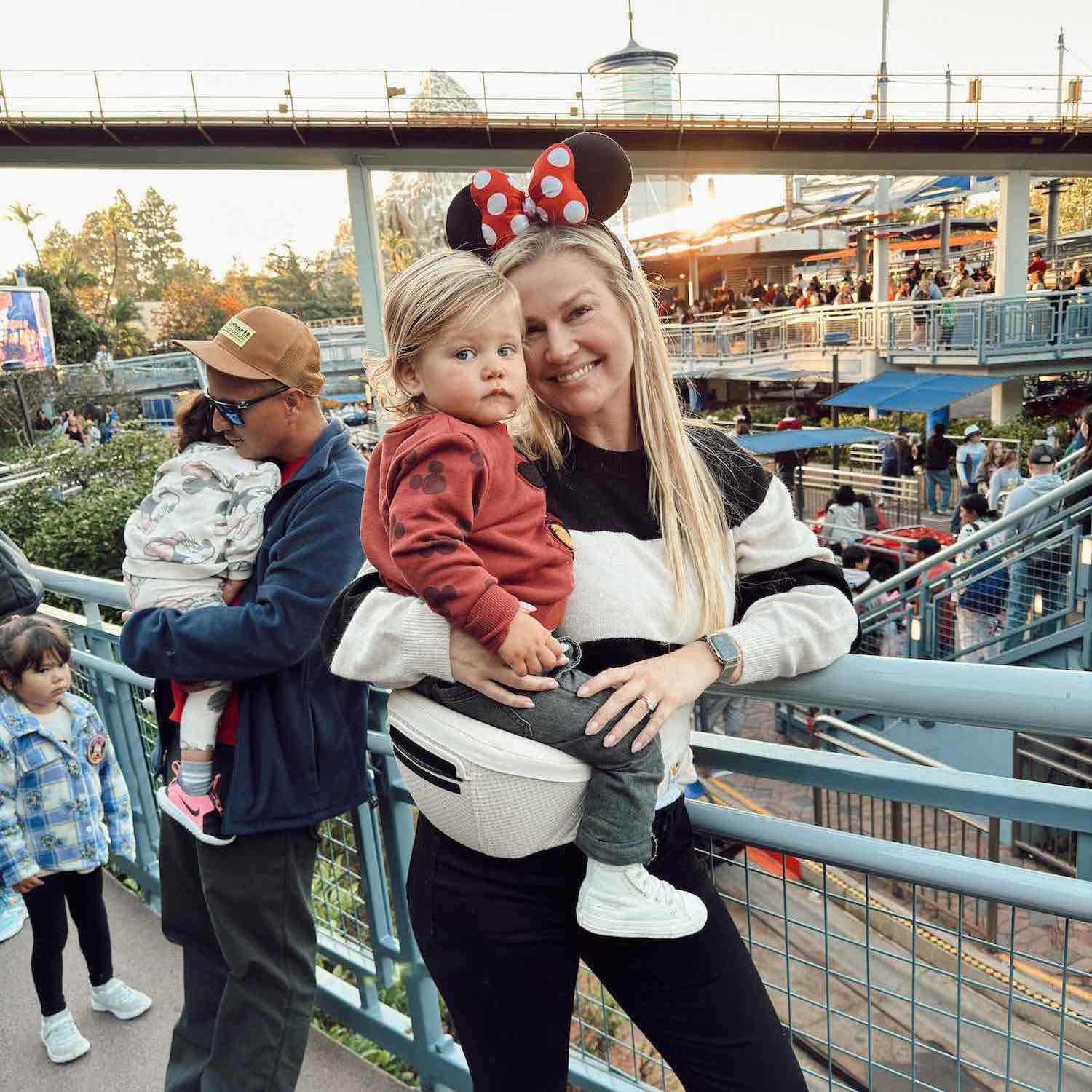 mom holding toddler in line at Disneyland while toddler sits on white hip carrier