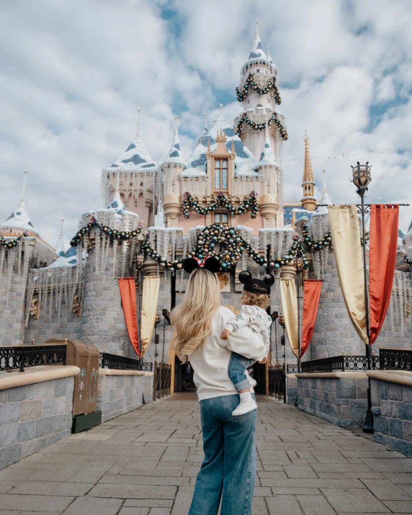 back of blonde woman with Minnie mouse ears holding blonde toddler boy in Mickey Mouse cap facing Sleeping Beauty Castle at Disneyland