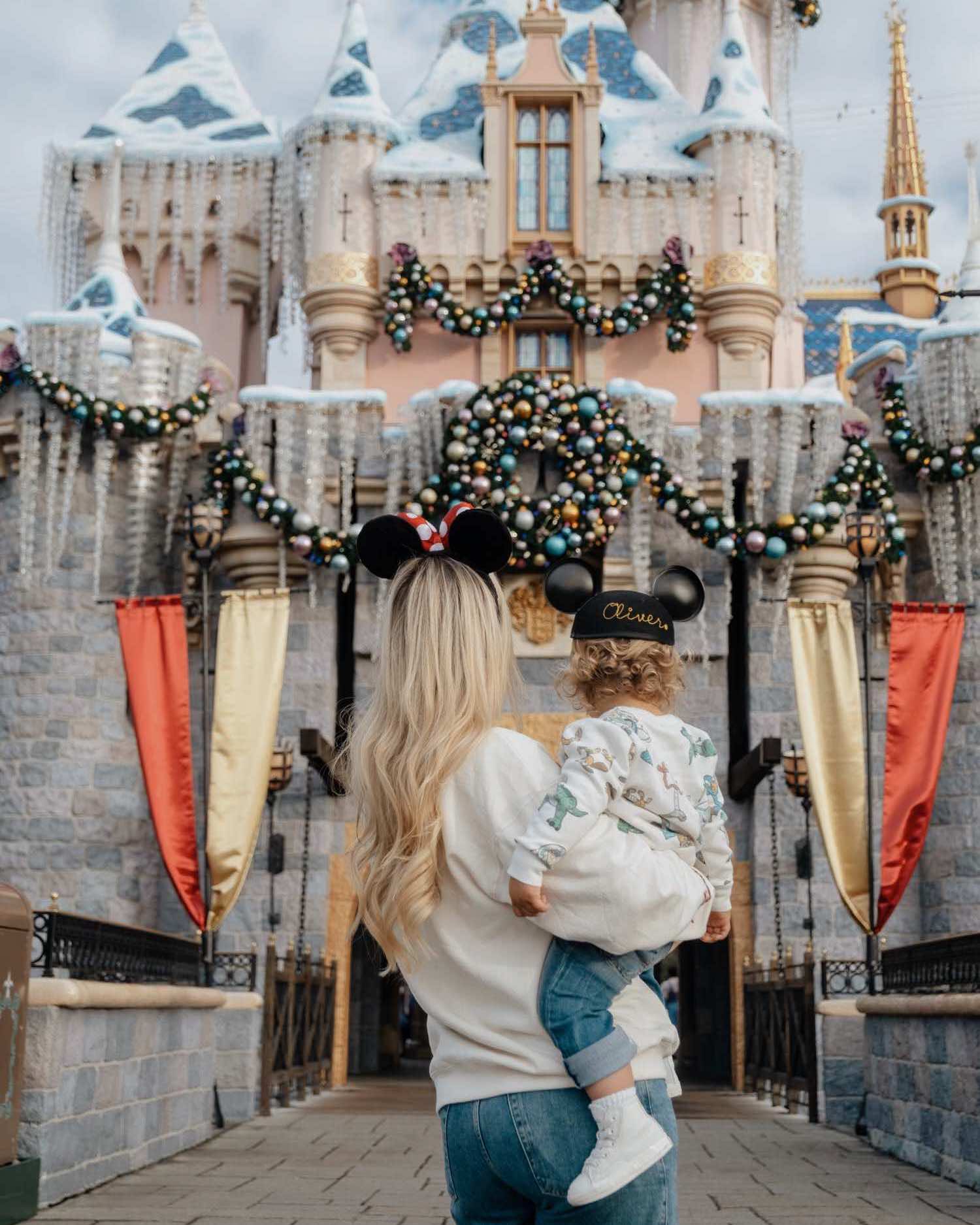 back of blonde woman with Minnie mouse ears holding blonde toddler boy in Mickey Mouse cap facing Sleeping Beauty Castle at Disneyland