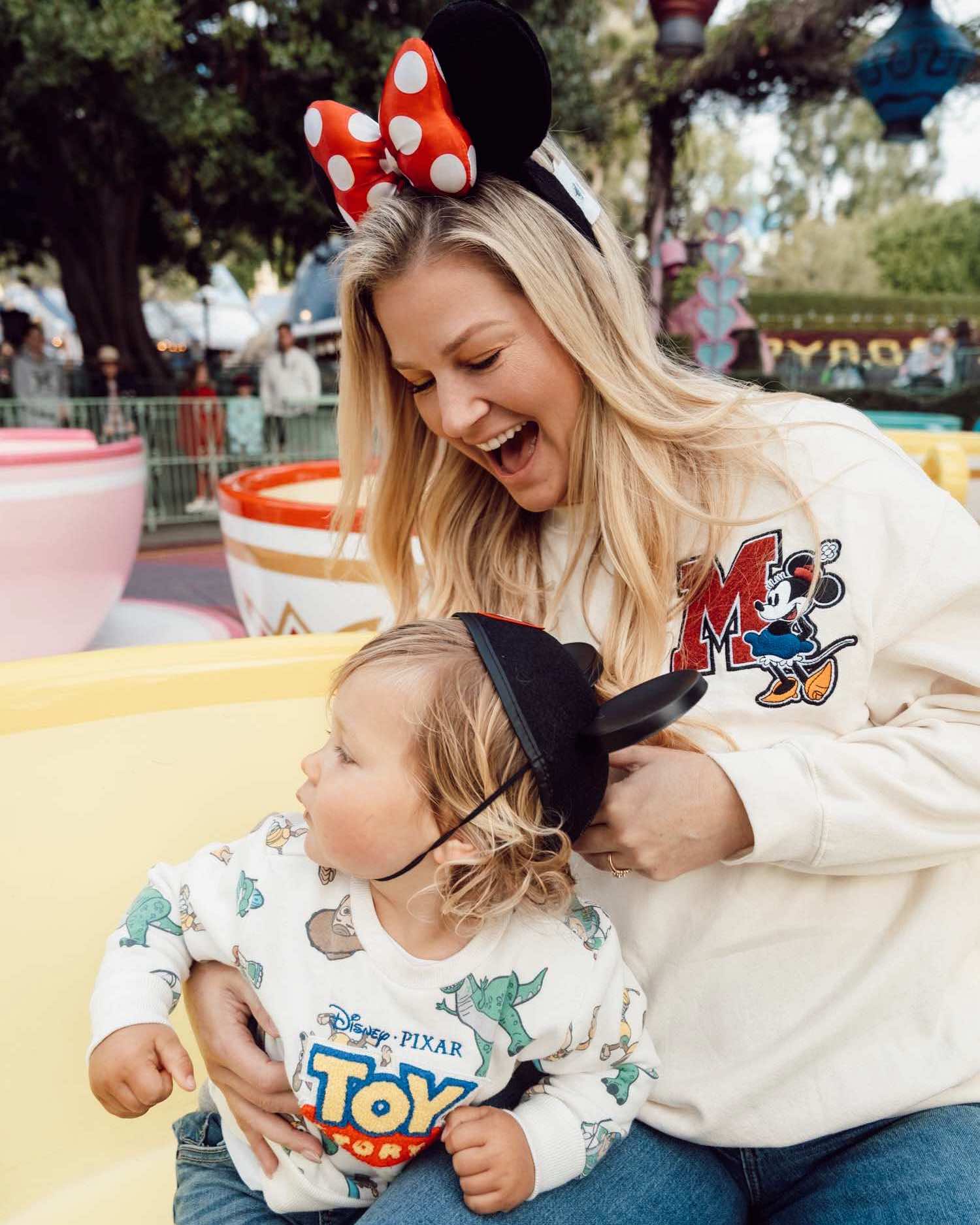 Blonde woman in Minnie sweater and Minnie ears looking down at toddler boy with Mickey ears as they ride yellow tea cup