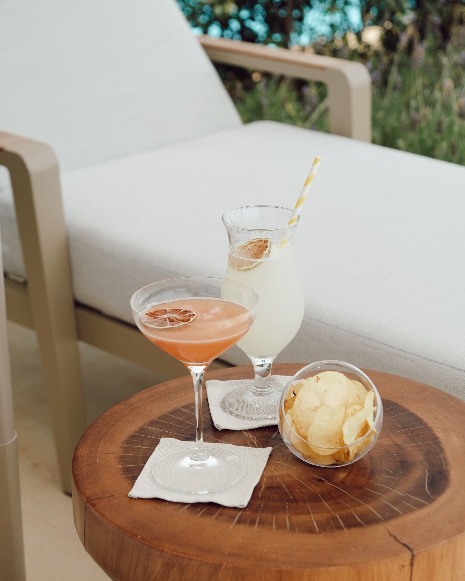 Cocktails with dried citrus garnish and bowl of chips on wooden side table next to beige lawn chair