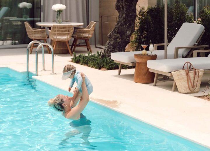 blonde woman in pool holding up baby in blue one piece suit overhead with luxury hotel lounge chairs in background