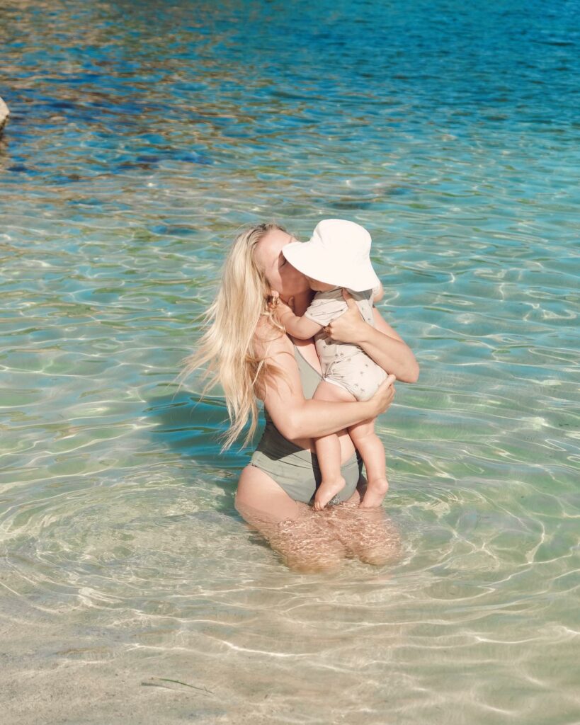 Close up of blonde woman in green one piece bathing suit sitting waist-deep in blue ocean holding baby with white sun hat