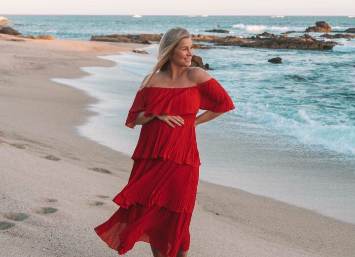 blonde woman in red tiered, off-the-shoulder dress with hand over baby bump with beach and ocean in the background