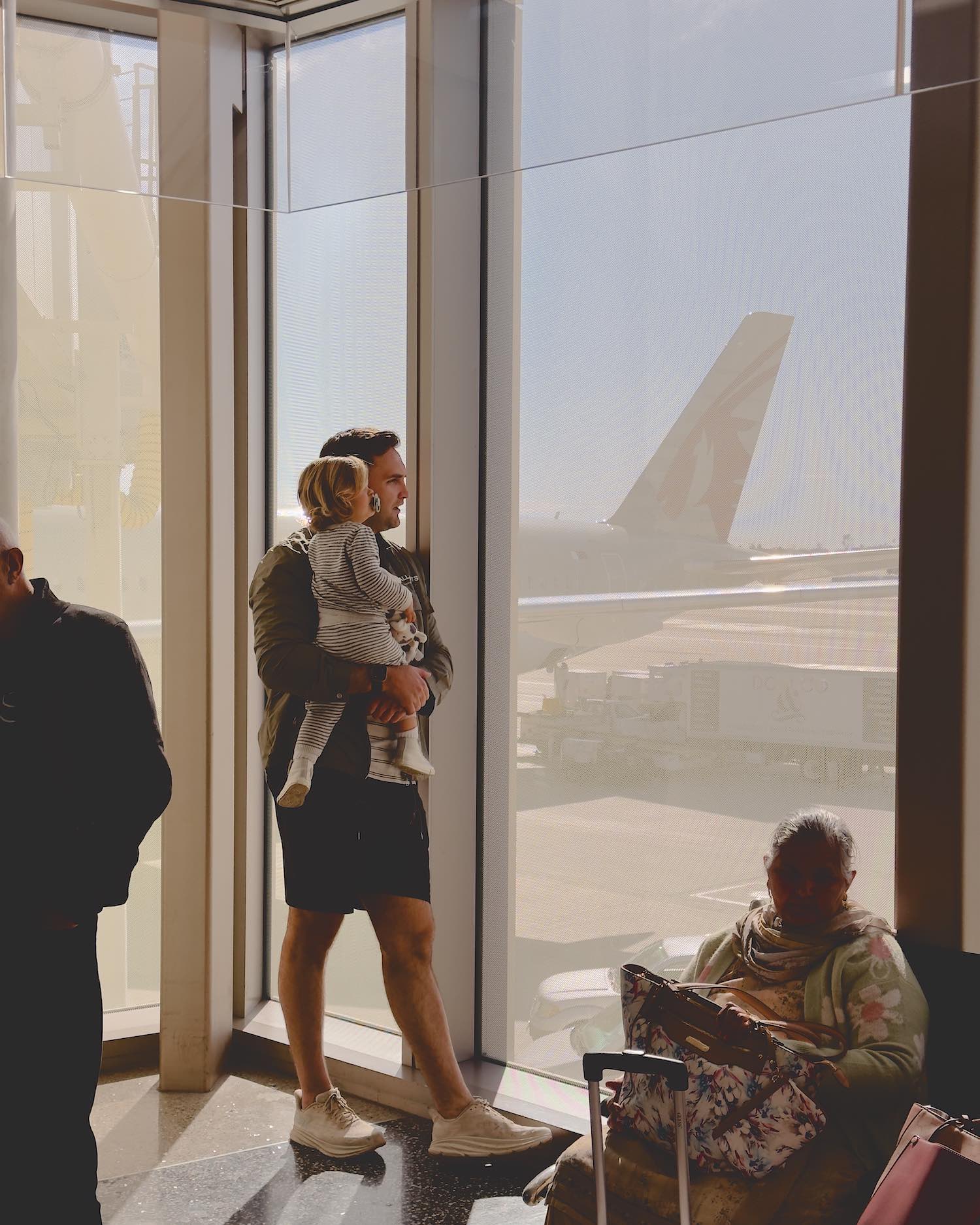 man in dark outfit holding toddler boy in pajamas looking out airport window at planes