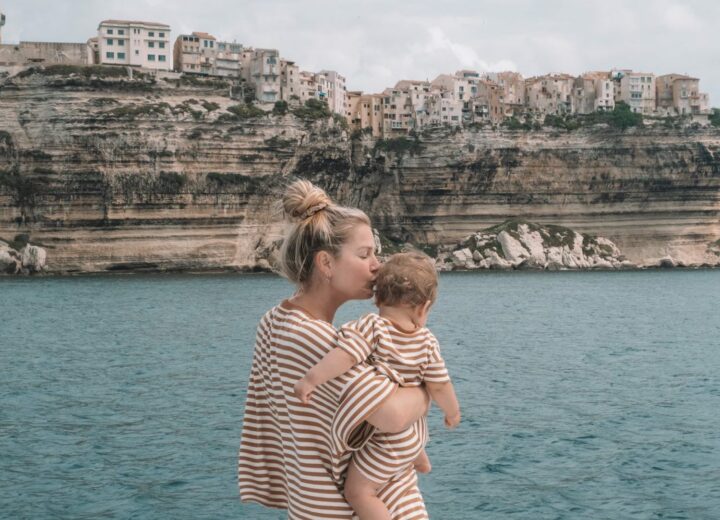 Blonde mom in stripes kissing baby in matching outfit overlooking the sea with homes on rocky cliffs in Italy