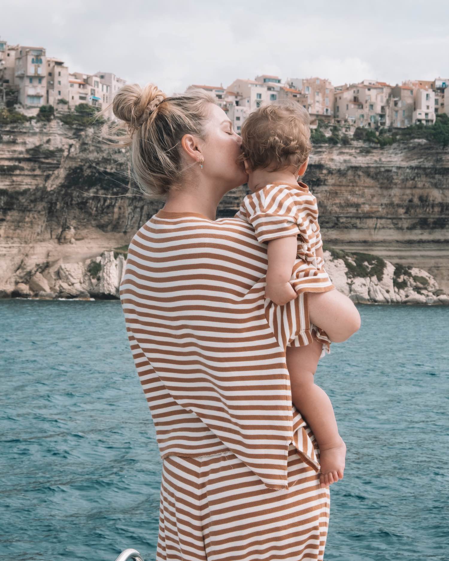 Close up image of back of blonde mom in stripes smiling at baby in matching outfit overlooking the sea with homes on rocky cliffs in Italy