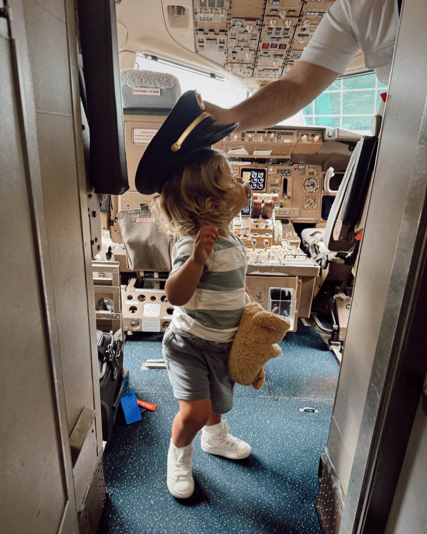 blonde toddler with pacifier in stripes with his teddy bear looking at out-of-frame pilot as pilot places captain's hat on toddler in cockpit