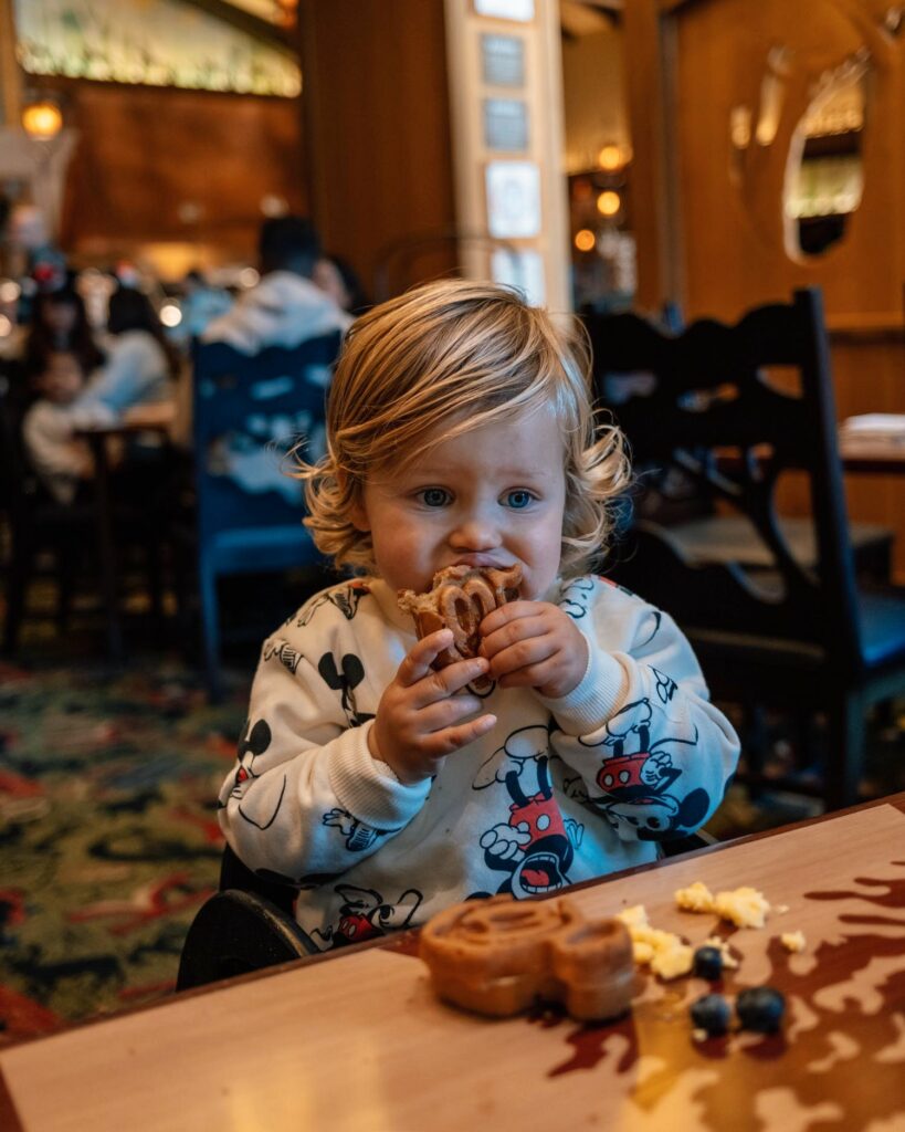 Blonde toddler boy in Mickey mouse sweatshirt eating Mickey waffles with waffles, eggs, and blueberries on table