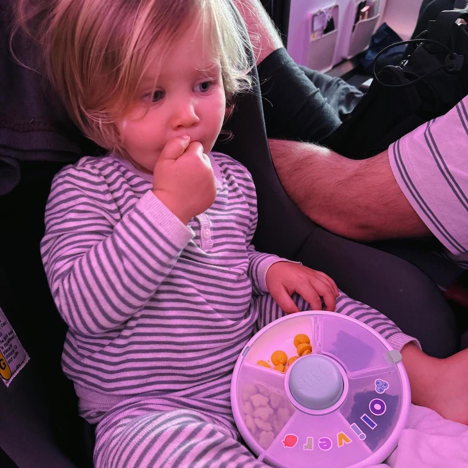 Blonde toddler in striped pajamas eating snacks out of snack box on airplane