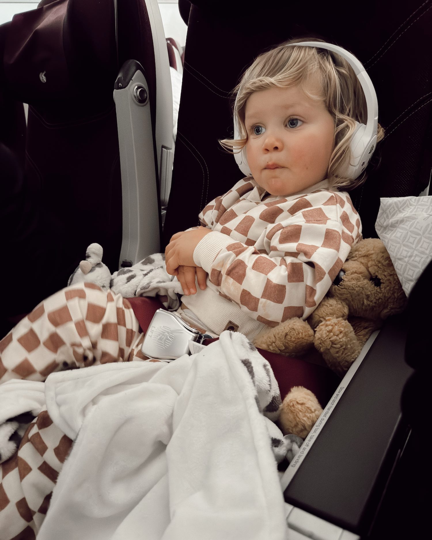 blonde toddler boy with headphones on and blanket sitting in airplane seat in brown checkered sweatsuit