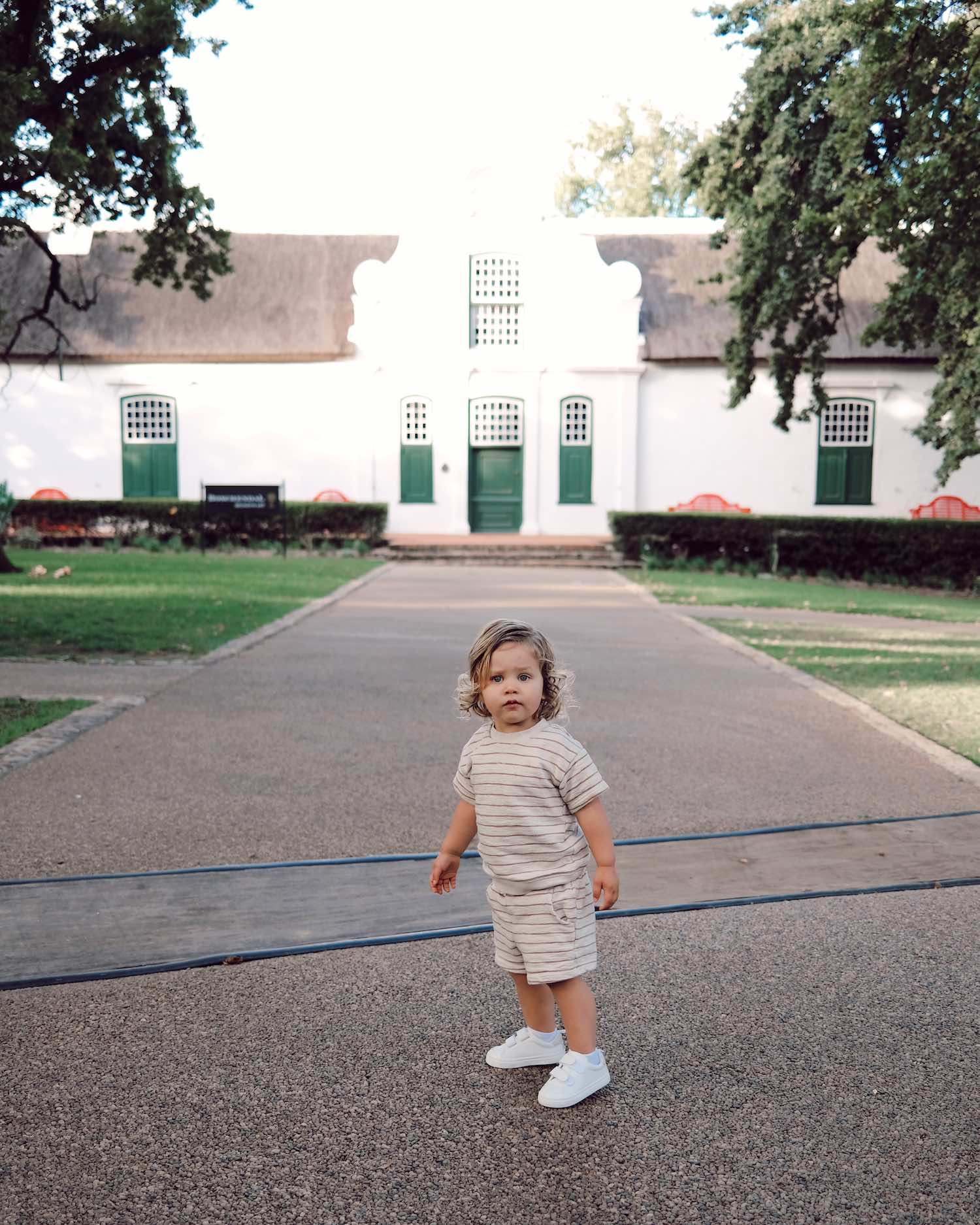 toddler boy in stripe short and tee set in front of large white house with trees on either side