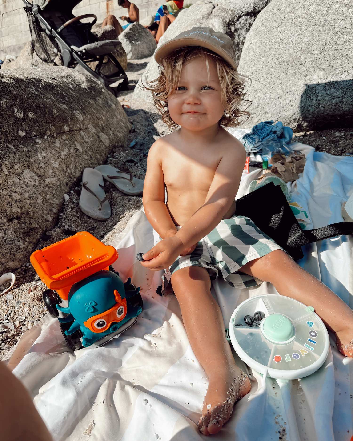 blonde boy in striped swim trunks and baseball cap with toy truck and snack box on the sandy beach