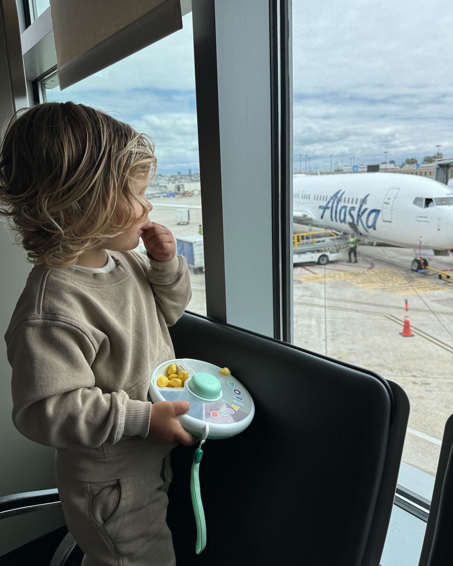 Toddler boy wearing beige sweats outfit looking out at plane from airport window as he eats snacks from box