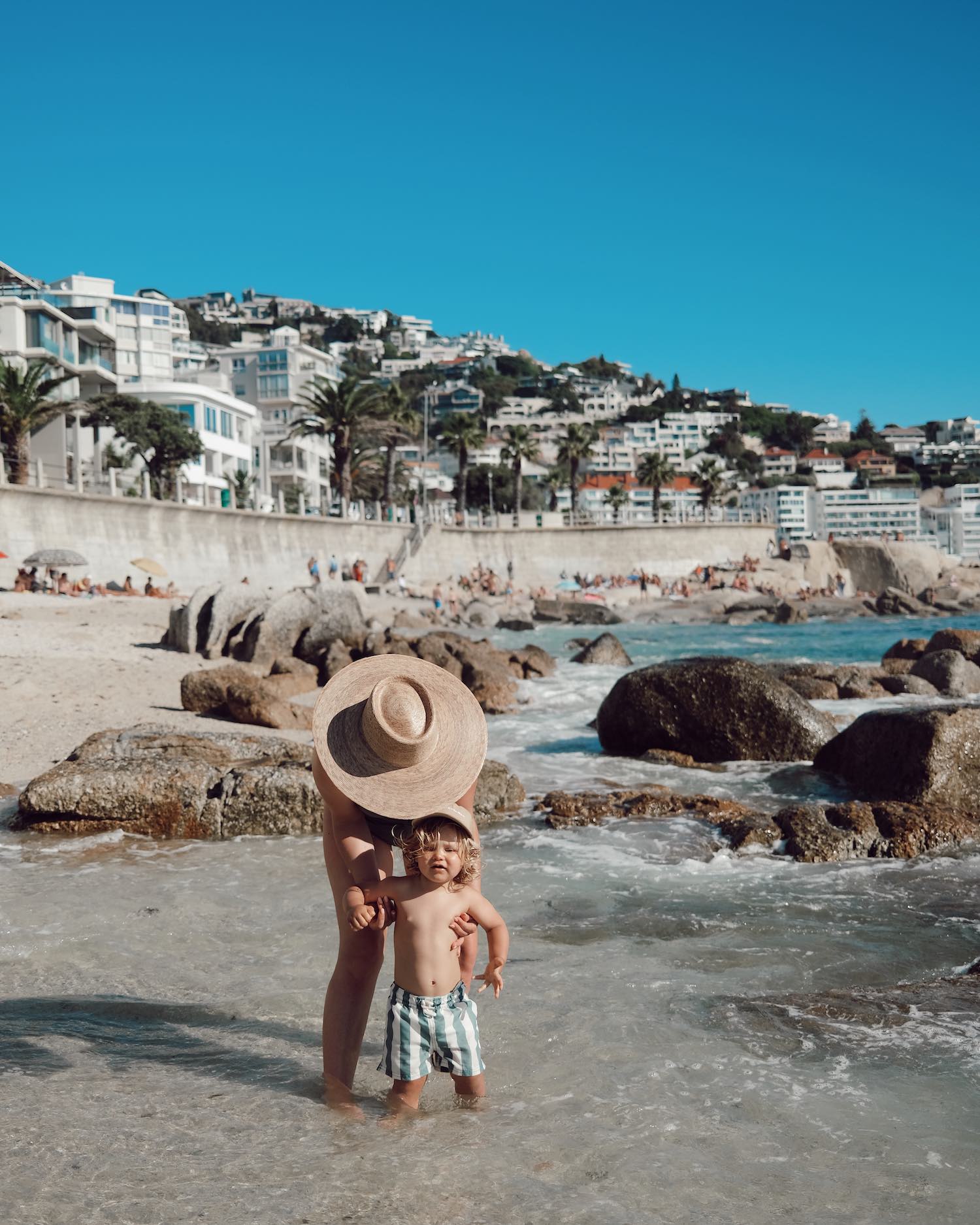 mom bending over in wide sun hat holding her toddler son in green striped swim trunks in the ocean