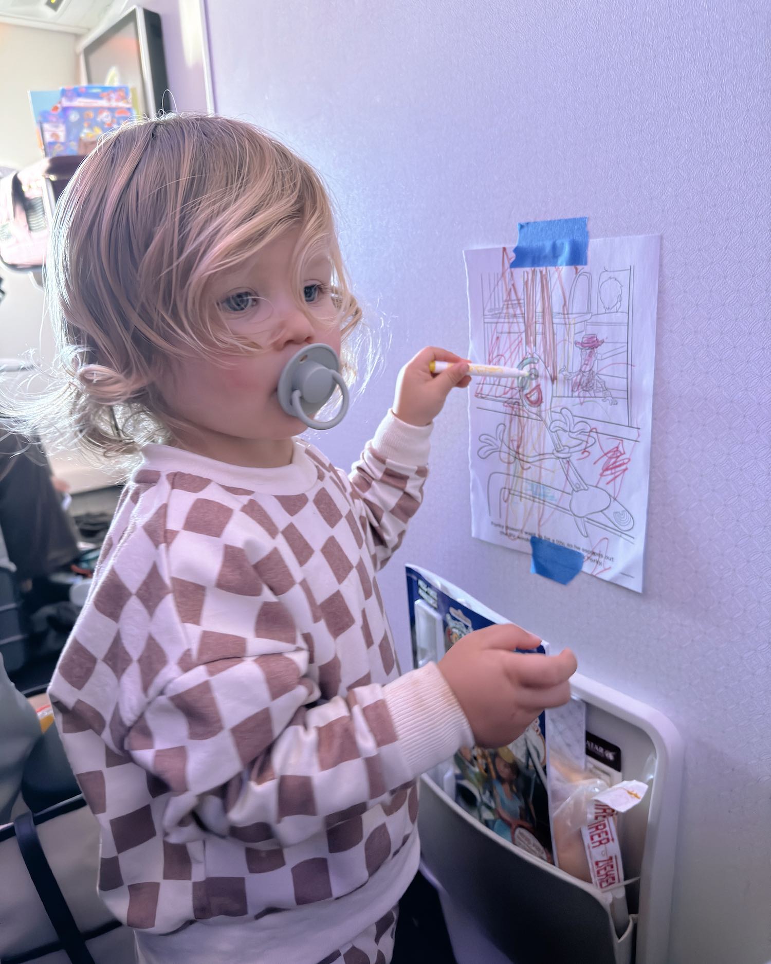 Toddler boy with pacifier wearing checked pajamas drawing on piece of paper that is taped to the wall of the plane bulk head