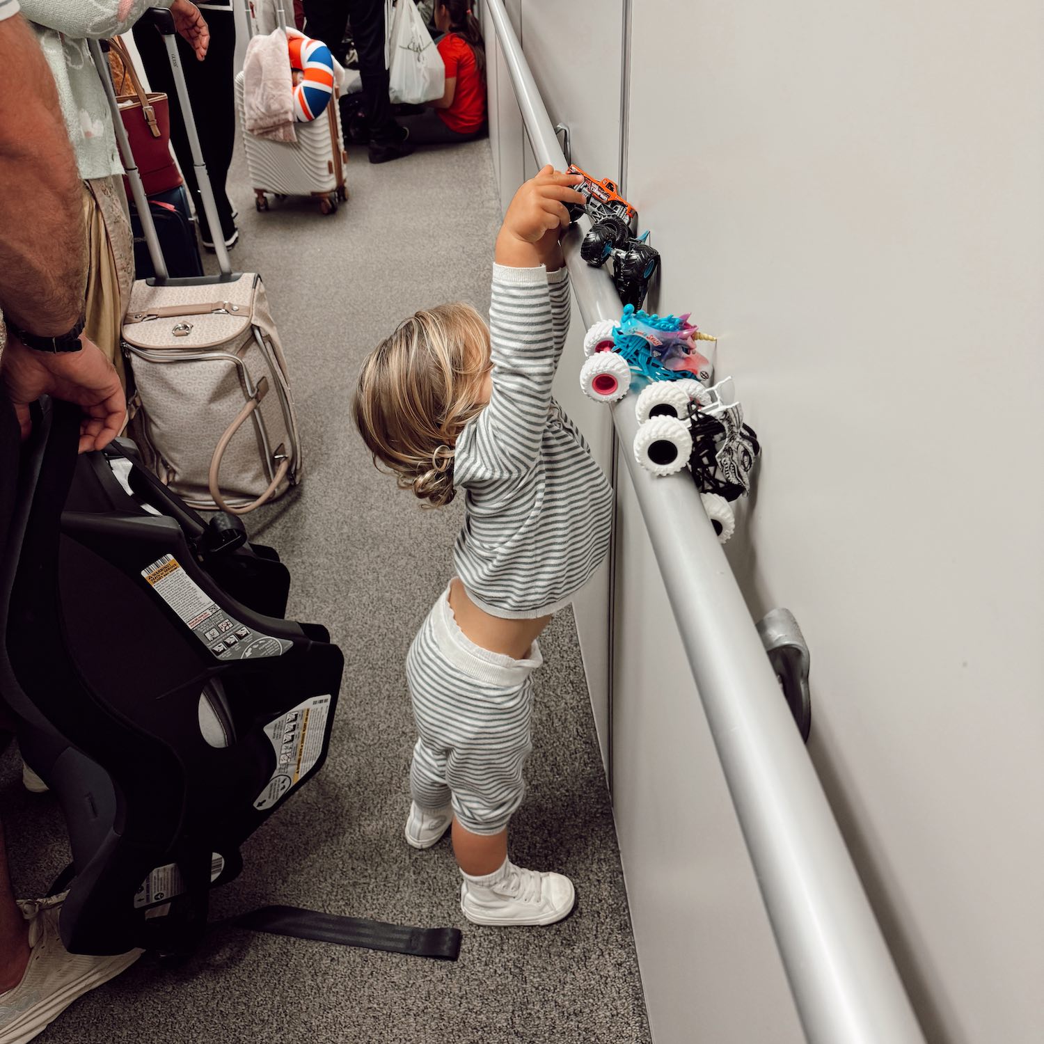 toddler in striped pajamas waiting in line to board airplane with luggage in foreground as he plays with truck toys