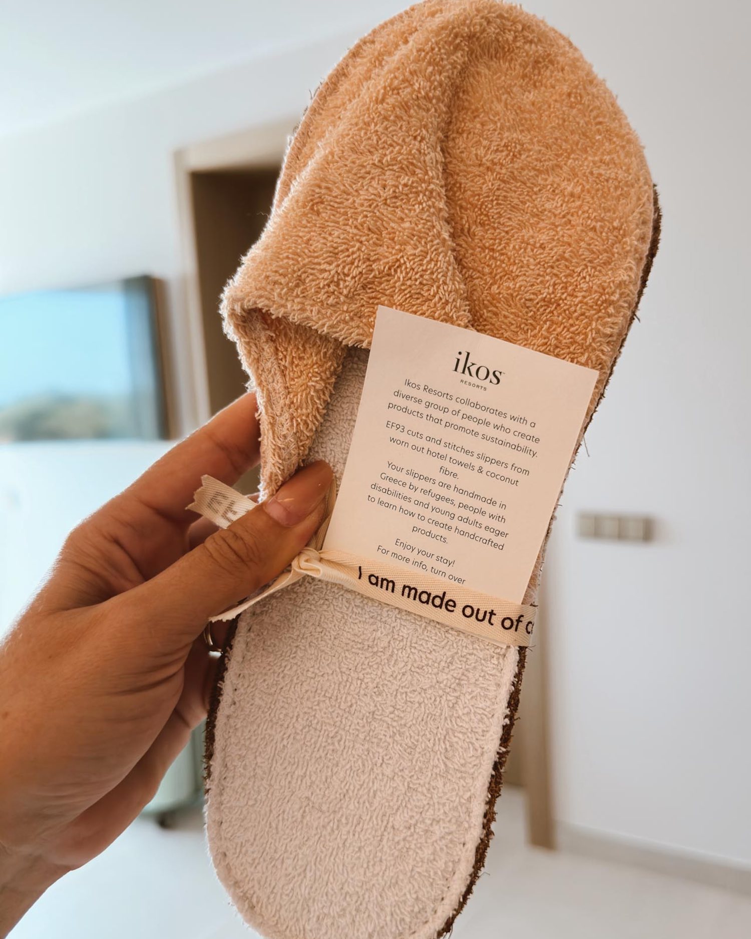 hand holding a terrycloth bath slipper with Ikos Porto Petro hotel logo on piece of paper with description tied with ribbon