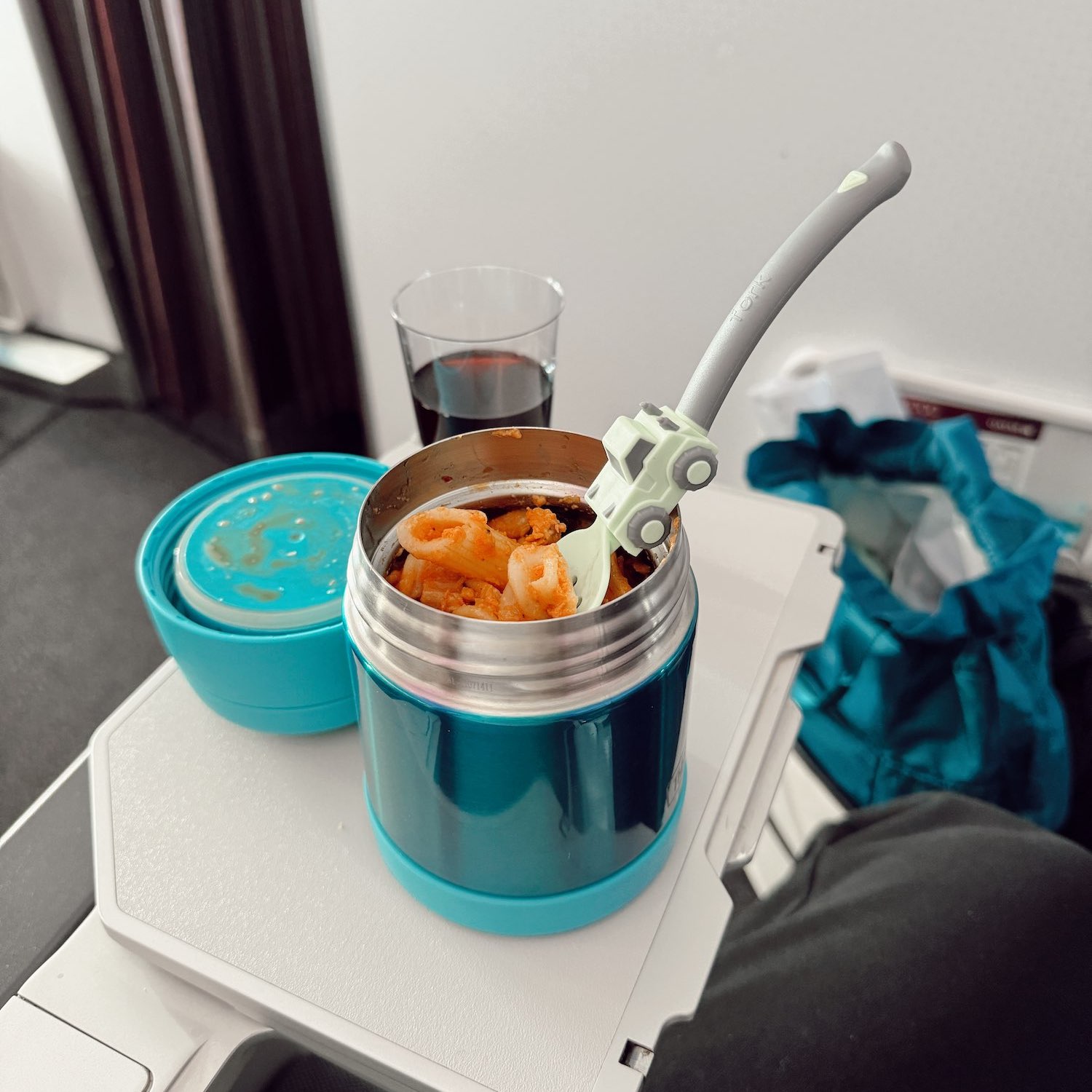 turquoise steel to-go cup with pre-made pasta and truck utensils next to a glass of wine on an airplane seat tray