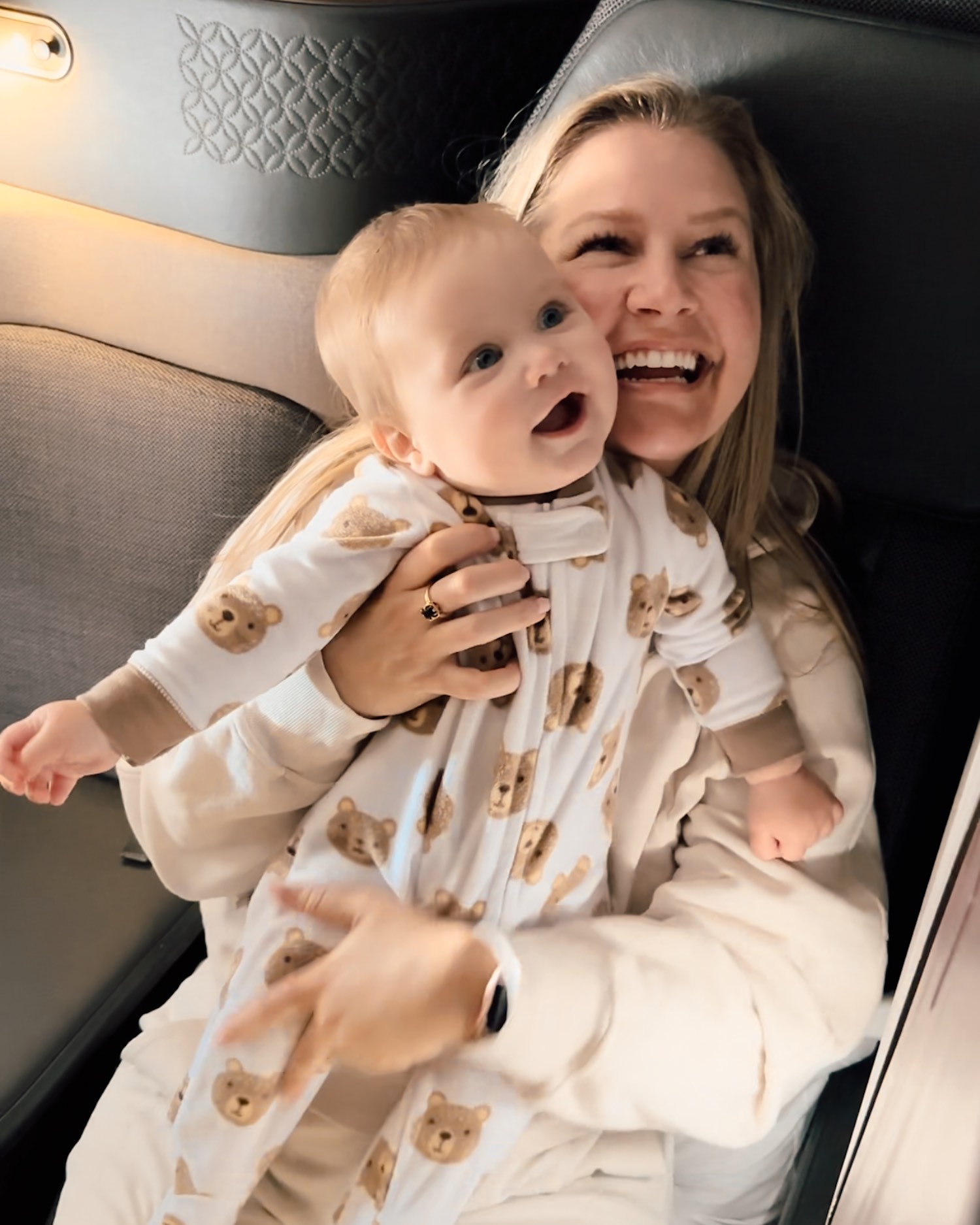 Blonde woman smiling off camera as she holds smiling baby looking in same direction in onesie as they sit in business class on airplane