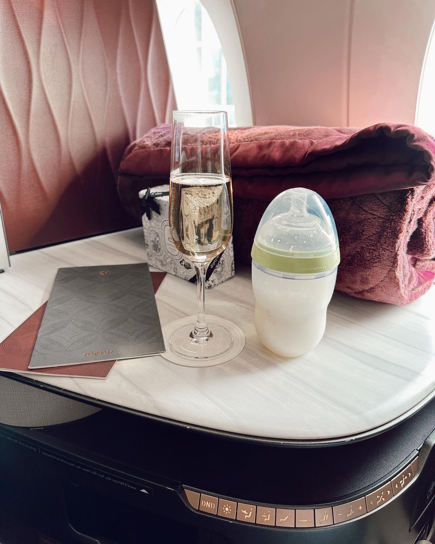 A small table on an airplane with a rolled up blanket, a half glass of champagne, and a baby milk bottle