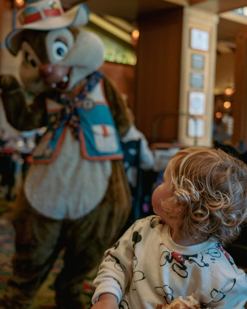 Toddler boy with curly blonde hair look back from table to see Dale Chipmunk waving hello at Disneyland