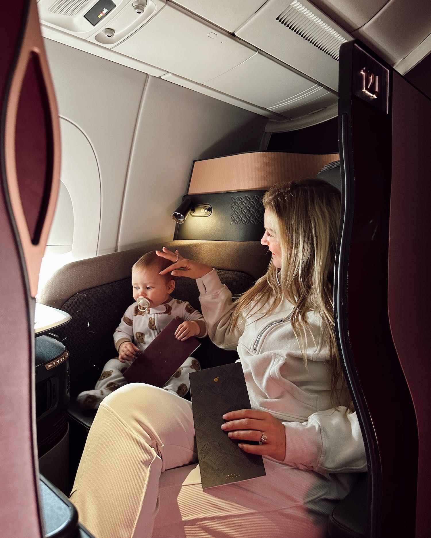 Woman enjoying window seat in Qatar QSuites looking lovingly at her baby as they both hold inflight menues with the sun shining in from the airplane window