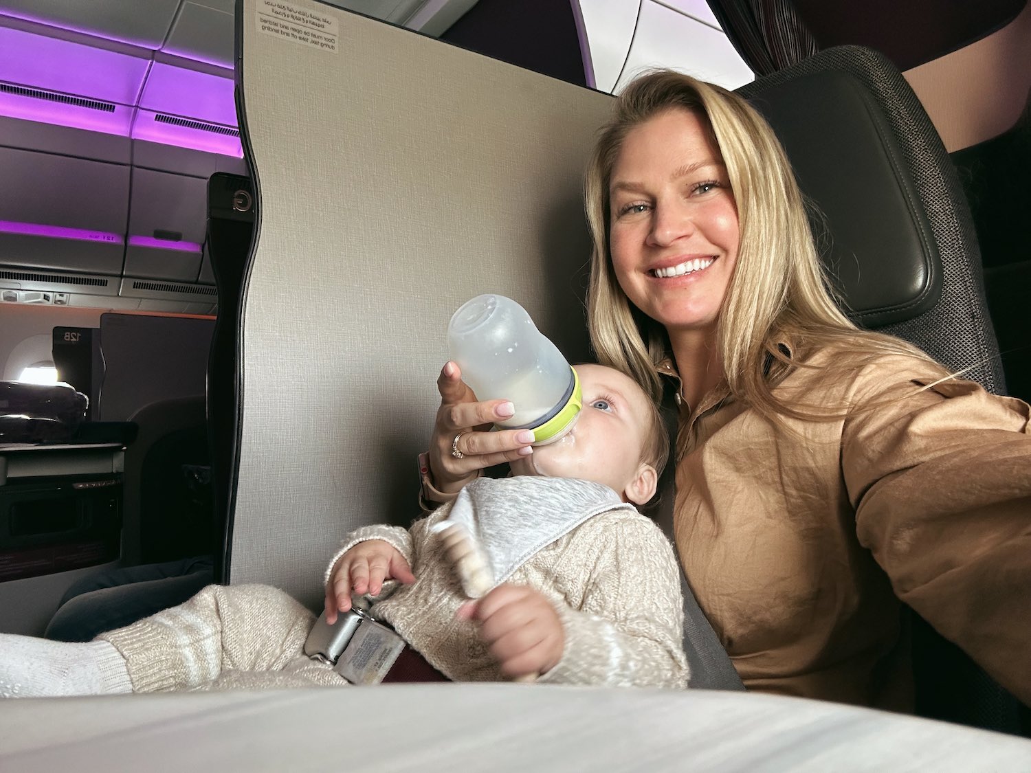 Blonde woman taking selfie in one hand with baby in another hand as she feeds him a bottle sitting in first class flight on Qatar Airways