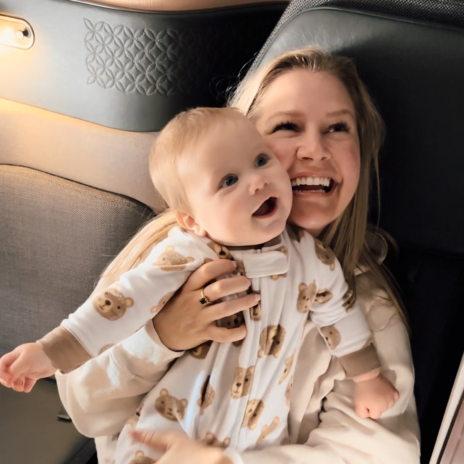 Blonde woman smiling off camera as she holds smiling baby looking in same direction in onesie as they sit in business class on airplane