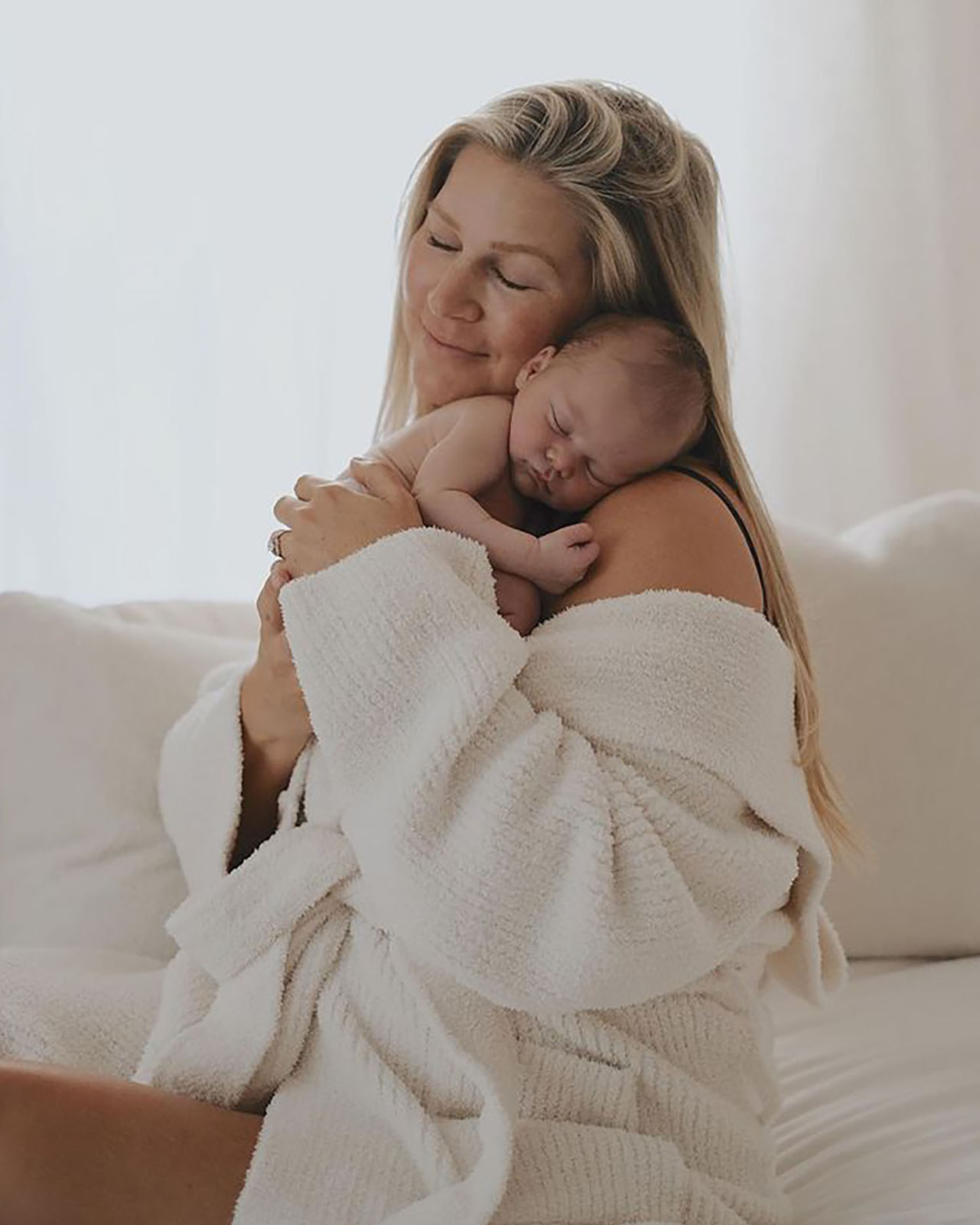 Blonde woman with her eyes closed and smiling wearing nothing but a white robe as she holds a small sleeping newborn close to her chest and on her shoulder