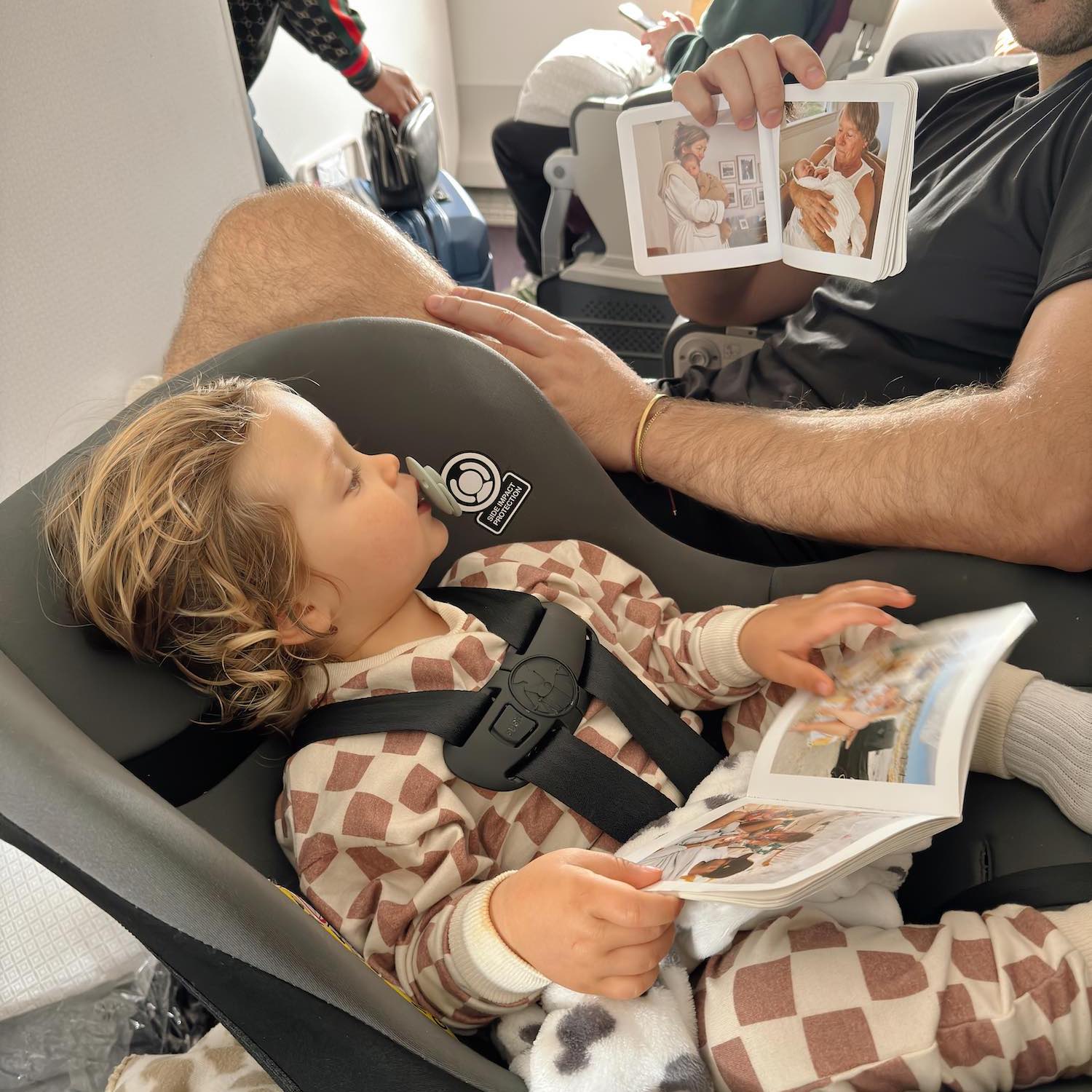 A man holding up a book to toddler also holding another book as he sits in carrier seat