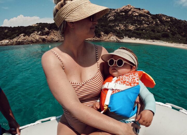 Go pro wide image of woman in striped swim suit and sun visor holding baby in sunglasses and life vest on dinghy over water