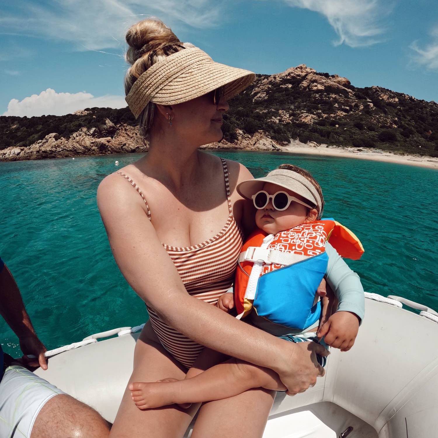 Go pro wide image of woman in striped swim suit and sun visor holding baby in sunglasses and life vest on dinghy over water