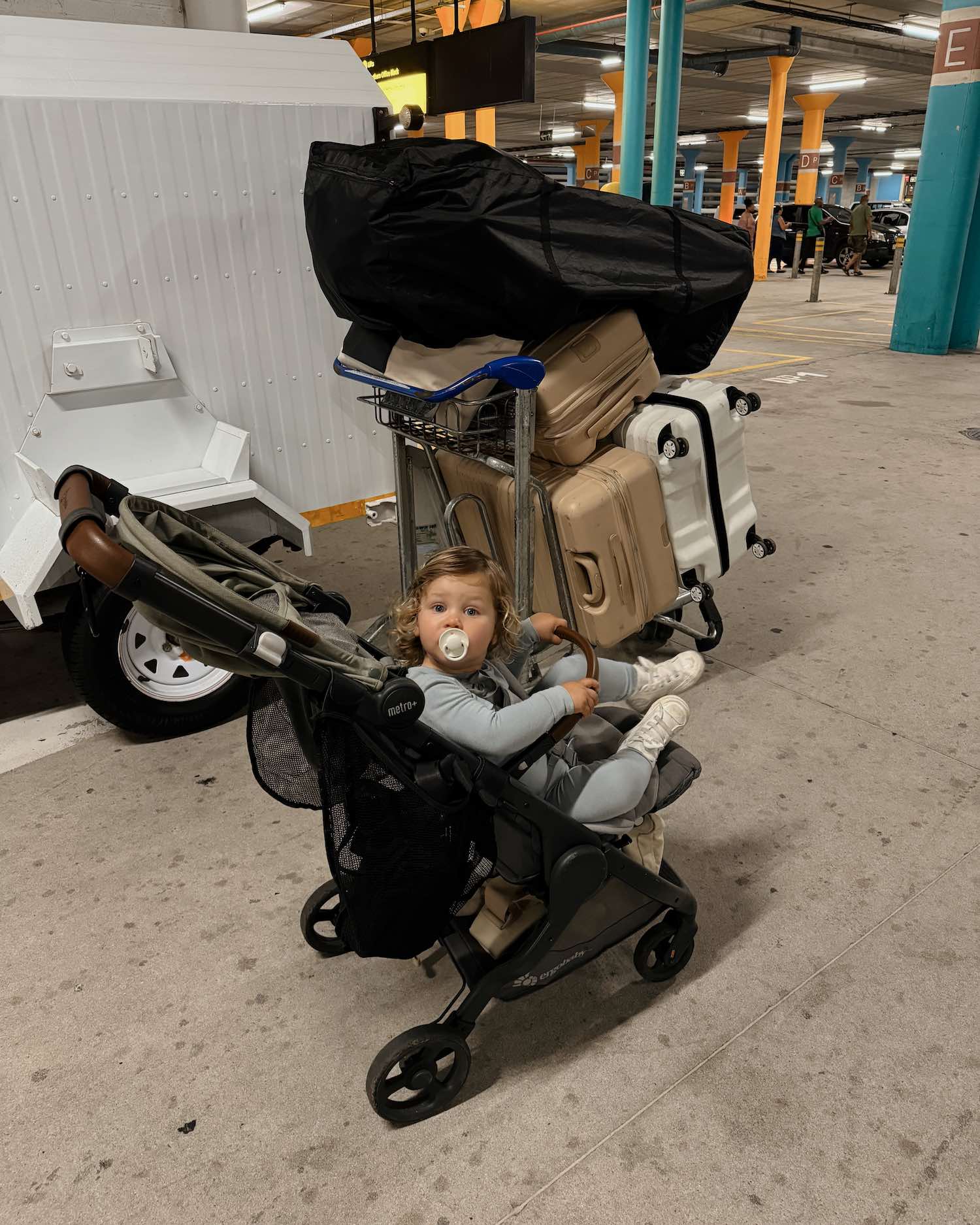 Little toddler boy in stroller with pacifier in mouth looking back at camera in airport with luggage around him