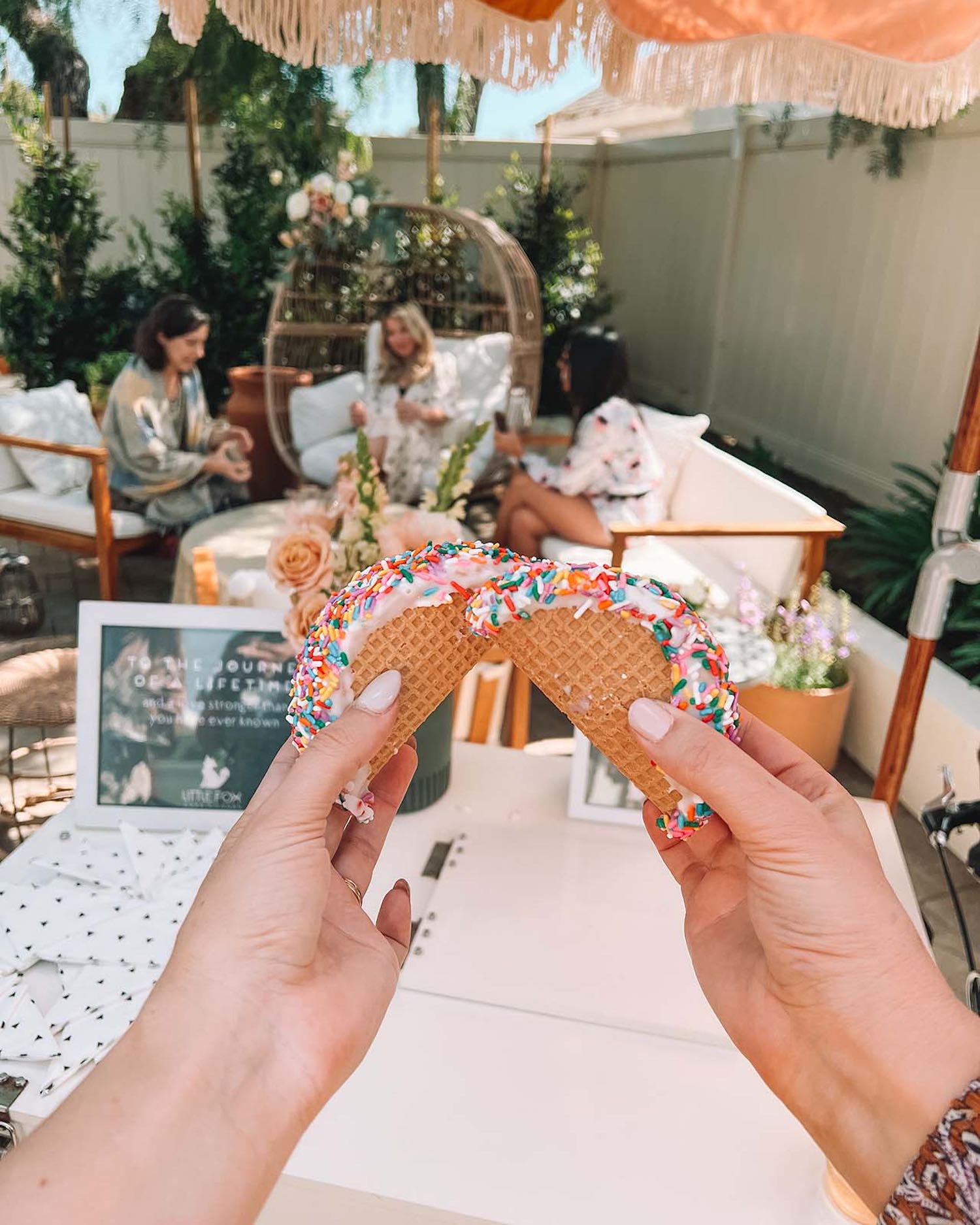 Two hands cheersing mini ice cream tacos with sprinkles with girls talking in the background