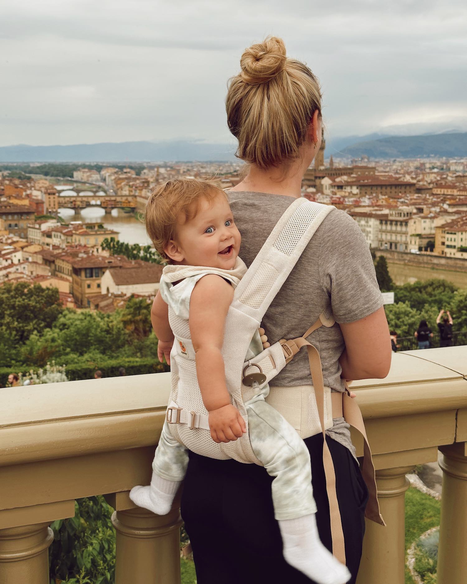 Woman with bun overlooking city from height behind banister with baby on her back looking back and smiling