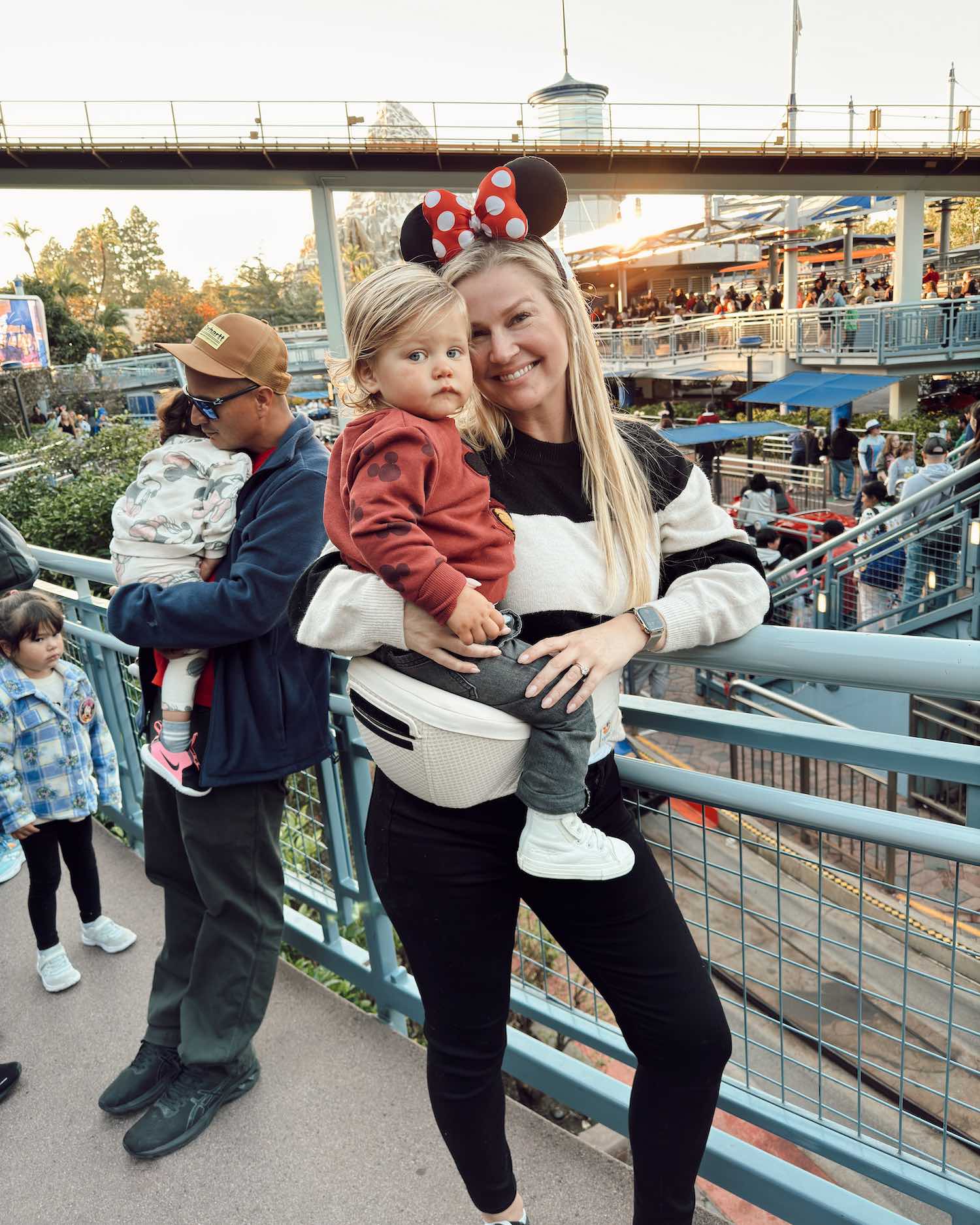 mom holding toddler in line at Disneyland while toddler sits on white hip carrier