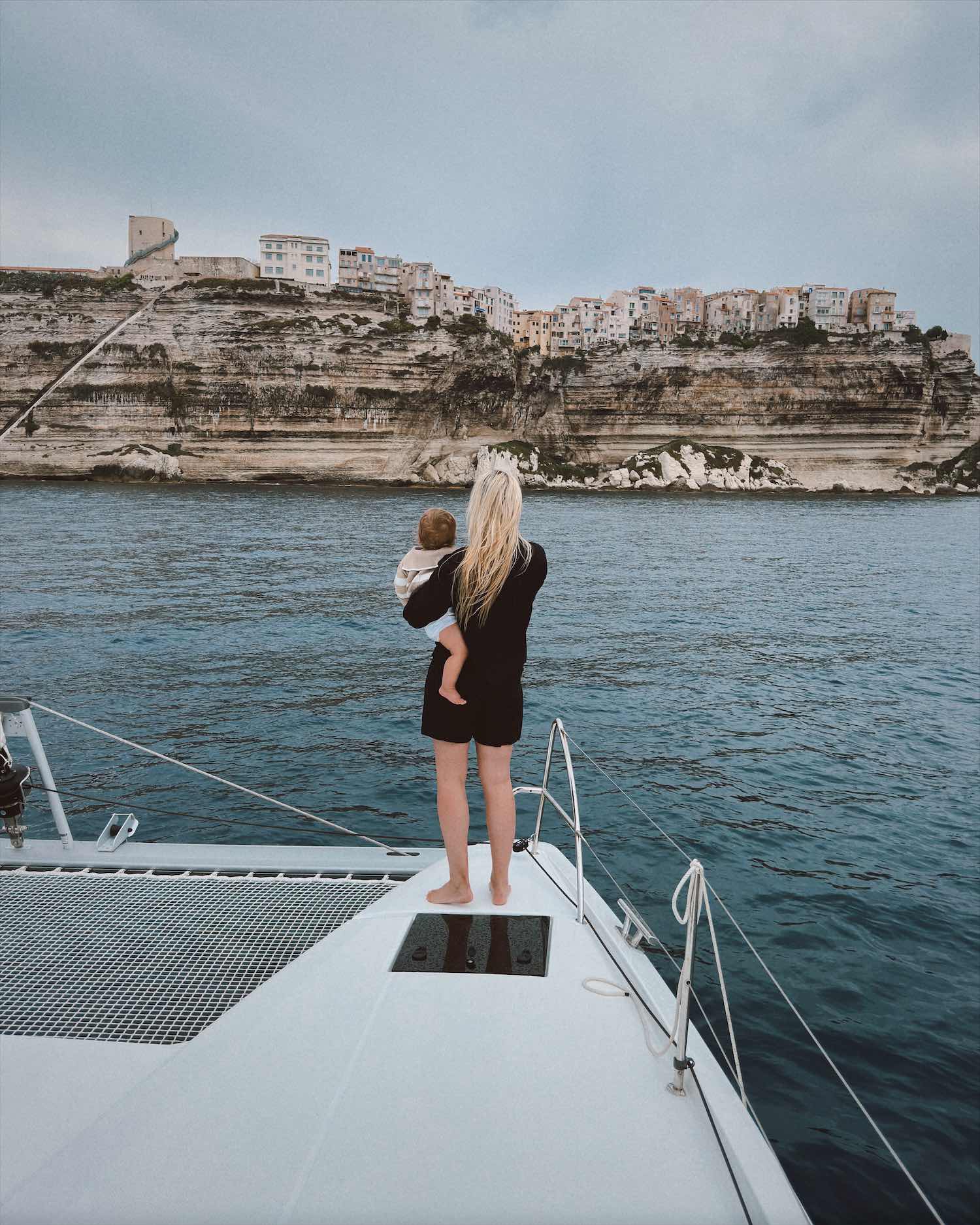 Woman in navy romper at the edge of catamaran over water holding baby with rocky cliffs and homes in the background