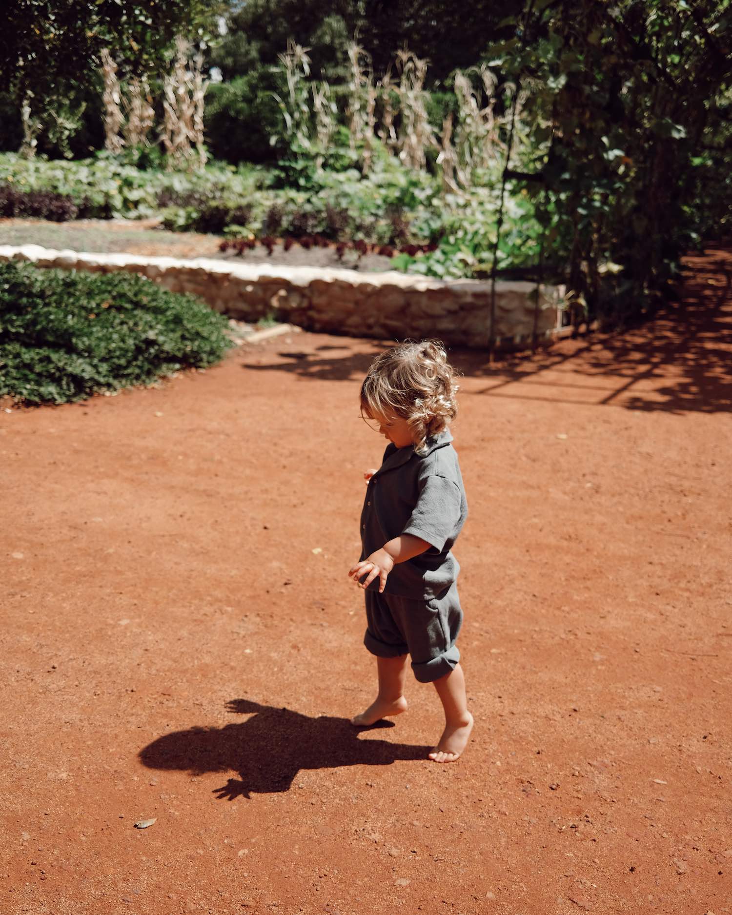 blonde toddler boy walking on his tip toes on orange dirt path with foliage in the background in matching cotton grey blue two piece