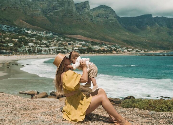 Woman in yellow dress and sun hat sitting on a rock lifting little baby up and kissing his forehead with vast ocean in the background and hills of South Africa in the background with blue skies and clouds behind