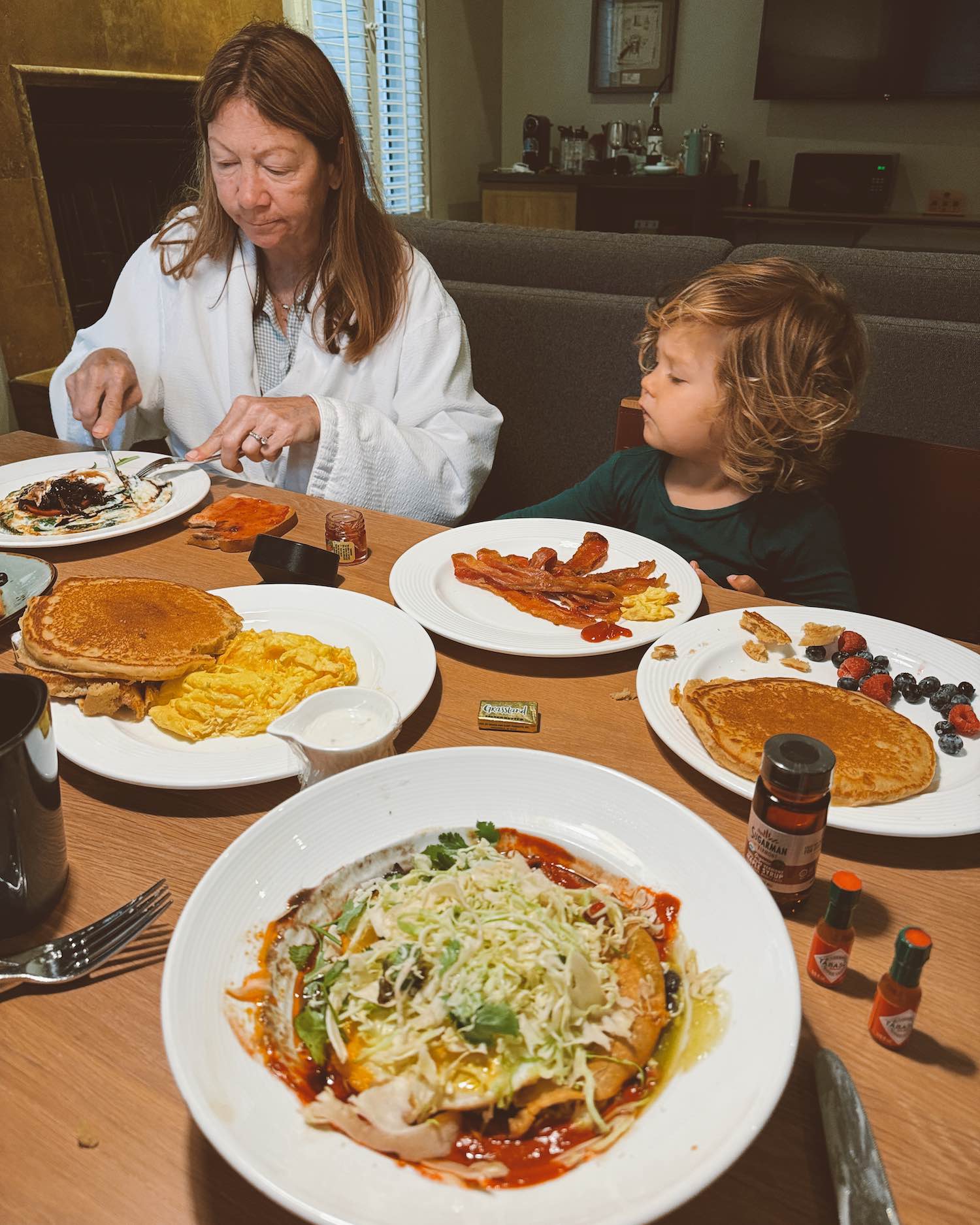 Woman in robe cutting into breakfast with little toddler boy looking at her as a table of breakfast food sits in front of them