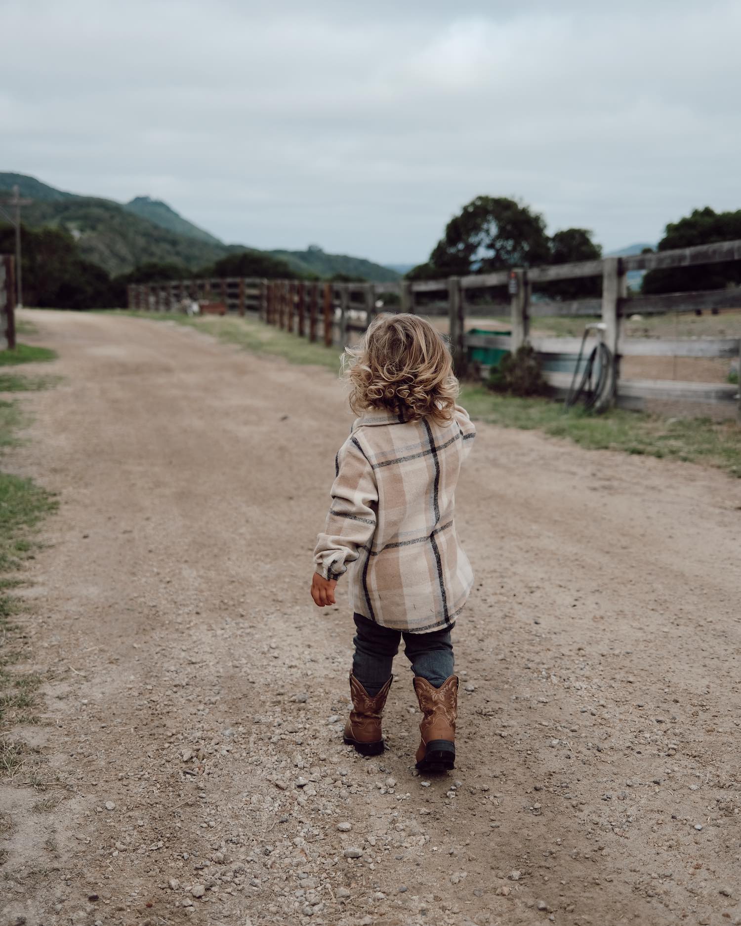 Little toddler boy with blonde girls walking along gravel pathway wearing beige flannel, jeans and brown cowboy boots