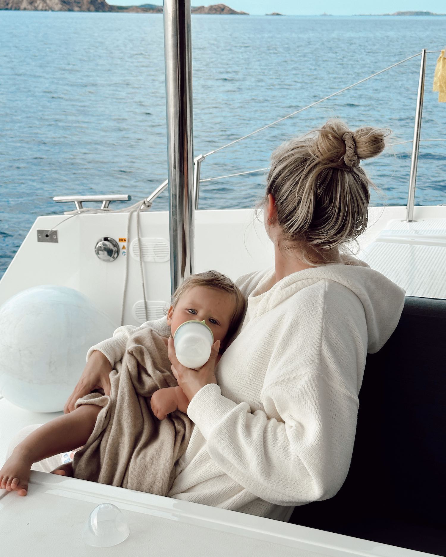 Woman with bun looking out at sea sitting on a boat with baby on her lap in a towel poncho as she feeds him baby bottle