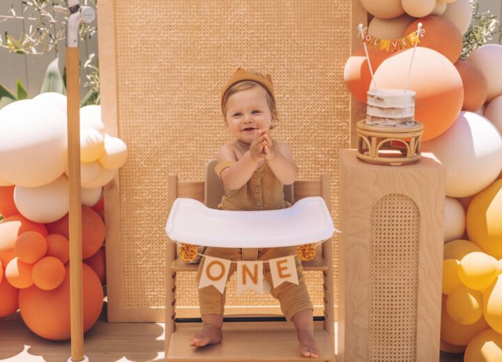 Little boy with crown on head as he sits in high chair with signs celebrating ONE year old clapping and smiling